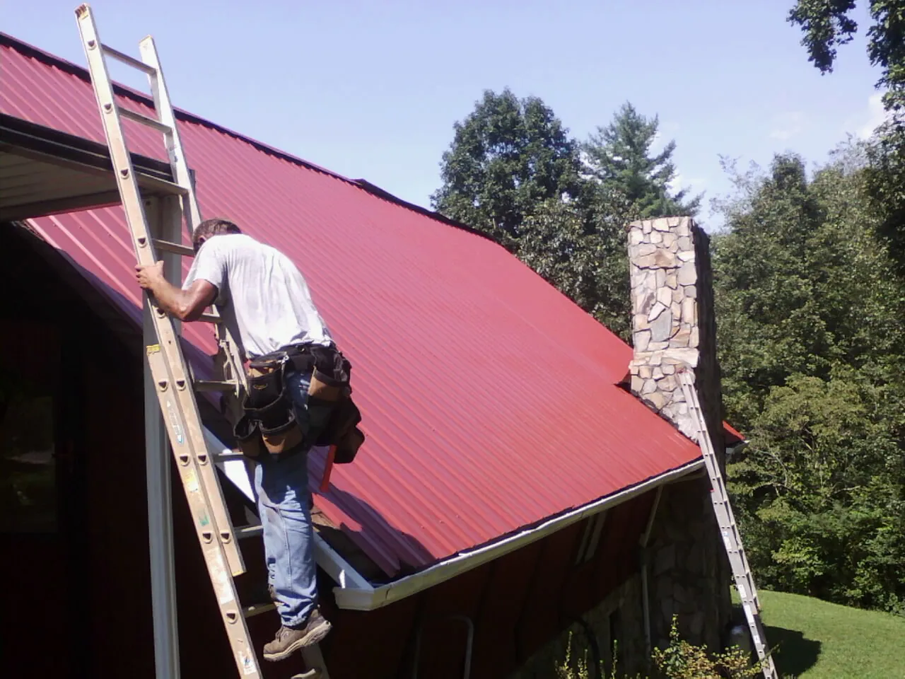 Roofer on a ladder beside a red metal roof and stone chimney — Soffit & Fascia Repair by Roofing Brothers in Chanhassen